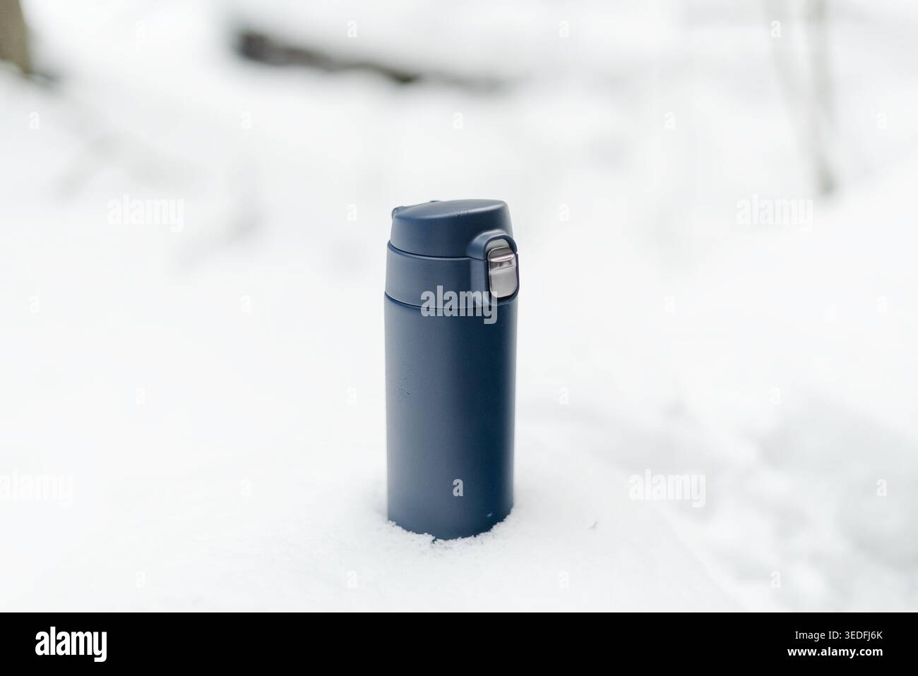 Dunkelblaue Thermoskanne steht vertikal im Neuschnee an einem eisigen Wintertag im Freien. Die isolierte Flasche erhebt sich über den eisigen Boden und ist bereit, heiße Getränke zu servieren. Unverzichtbare Ausrüstung für Winterwanderungen Stockfoto
