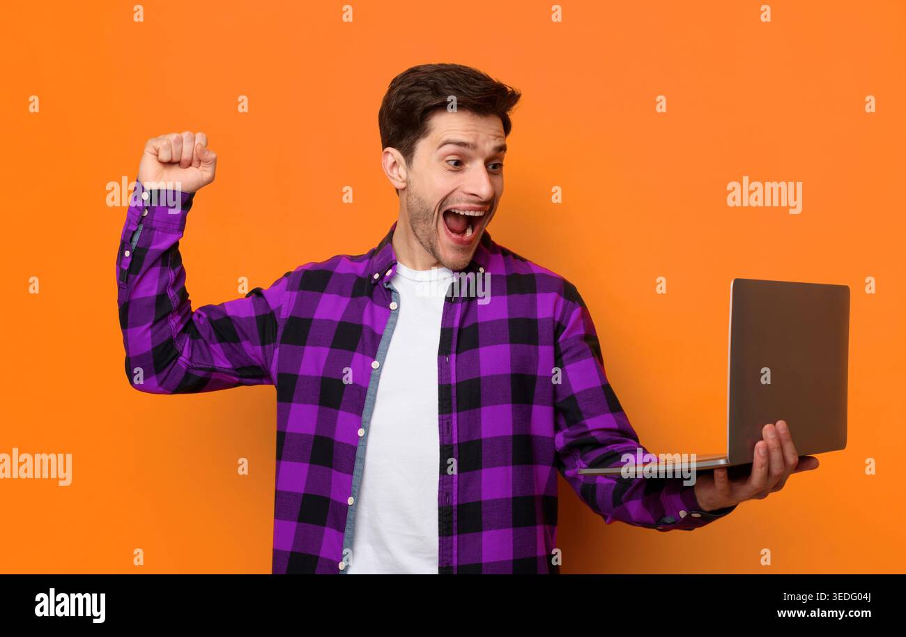 Junger Mann feiert, während er einen Laptop vor einer orangefarbenen Wand hält Stockfoto