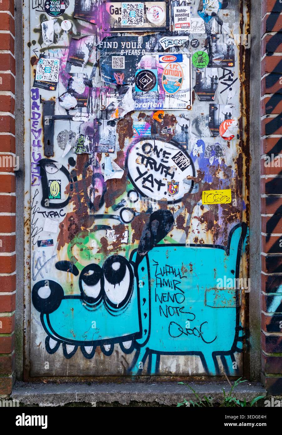 Graffiti-überzogene Metalltür mit einem türkisfarbenen Hund im Comic-Stil und mehrschichtigen Street-Art-Aufklebern in Berlin, Deutschland. Stockfoto