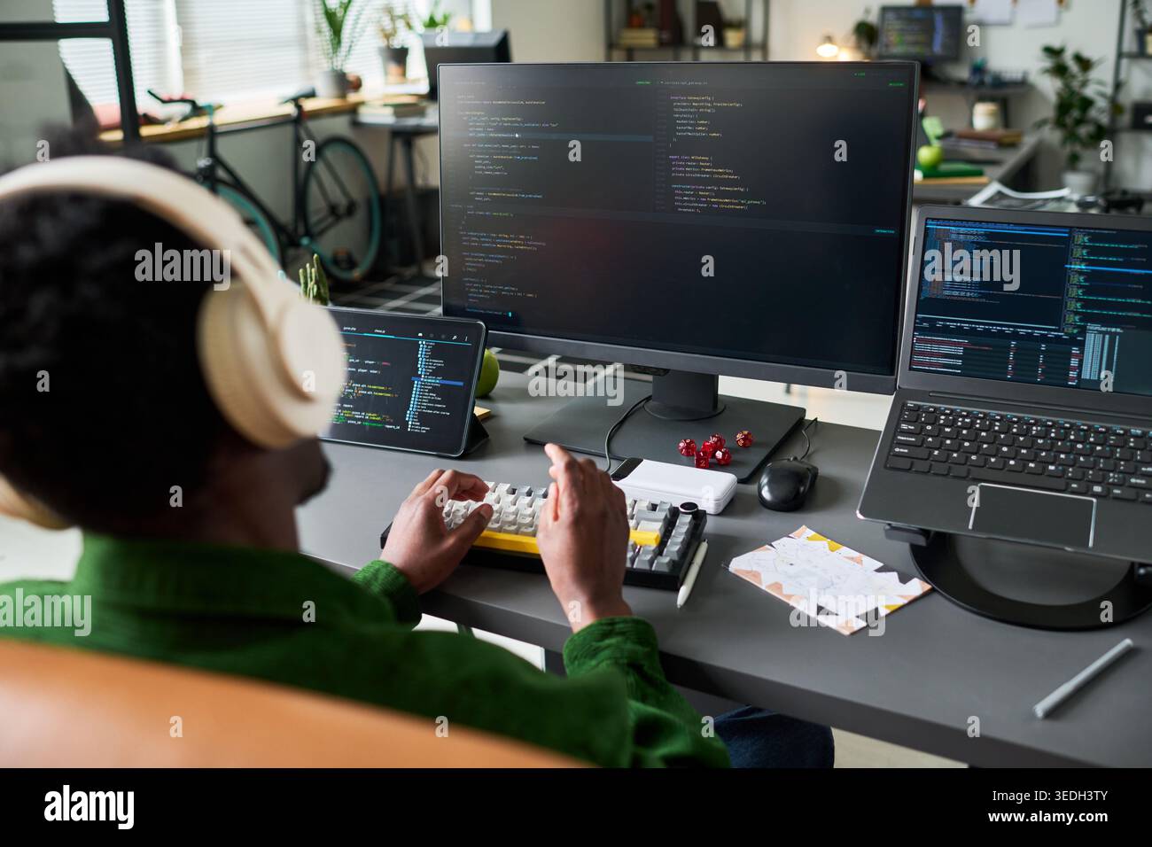 Afroamerikanischer junger erwachsener Mann, der an einer Workstation mit mehreren Bildschirmen codiert, Kopfhörer trägt und auf dem Desktop-Setup eine Tippsoftware verwendet. Ideal für Remote-Arbeiten, Entwicklerkarriere und technische Produktivität Stockfoto
