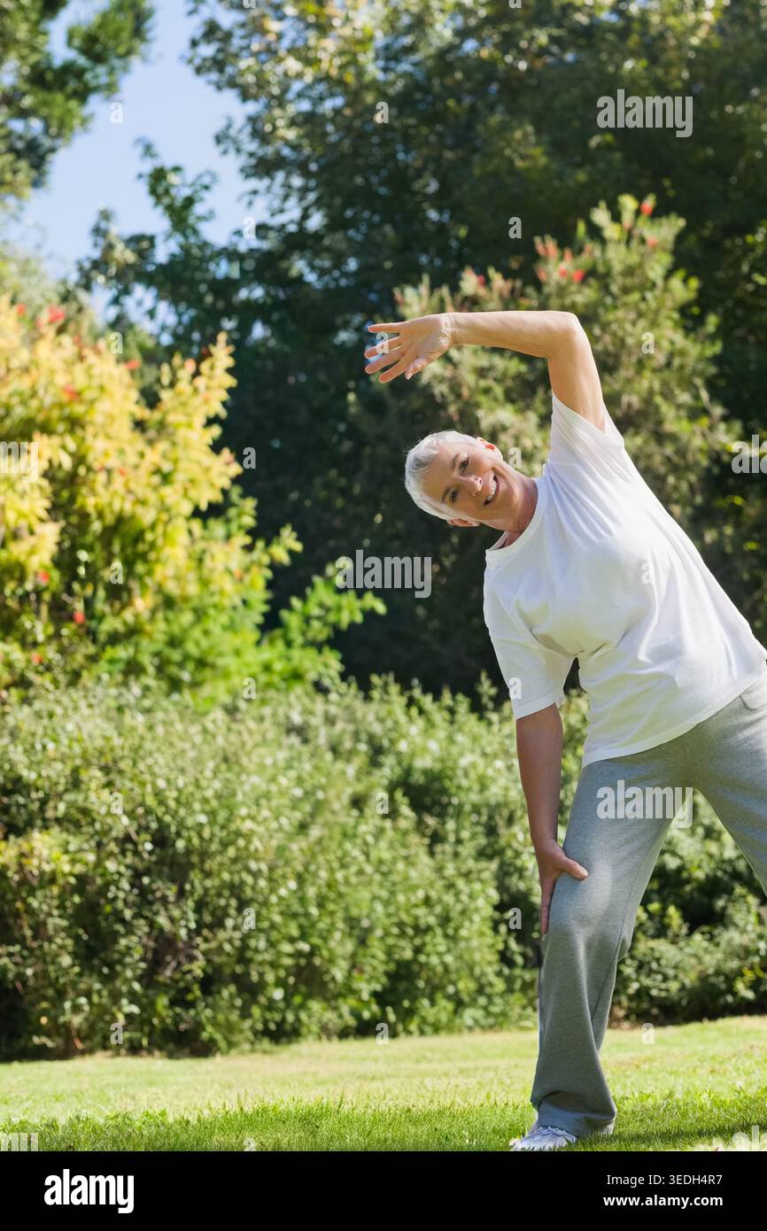 Seniorin trainiert seitliche Dehnung auf grasbewachsenem Rasen mit Sträuchern, weißem T-Stück grauen Schweißausbrüchen, Kopierraum. Ältere Menschen, Outdoor, Fitness, Wellness, Natur, Garde Stockfoto