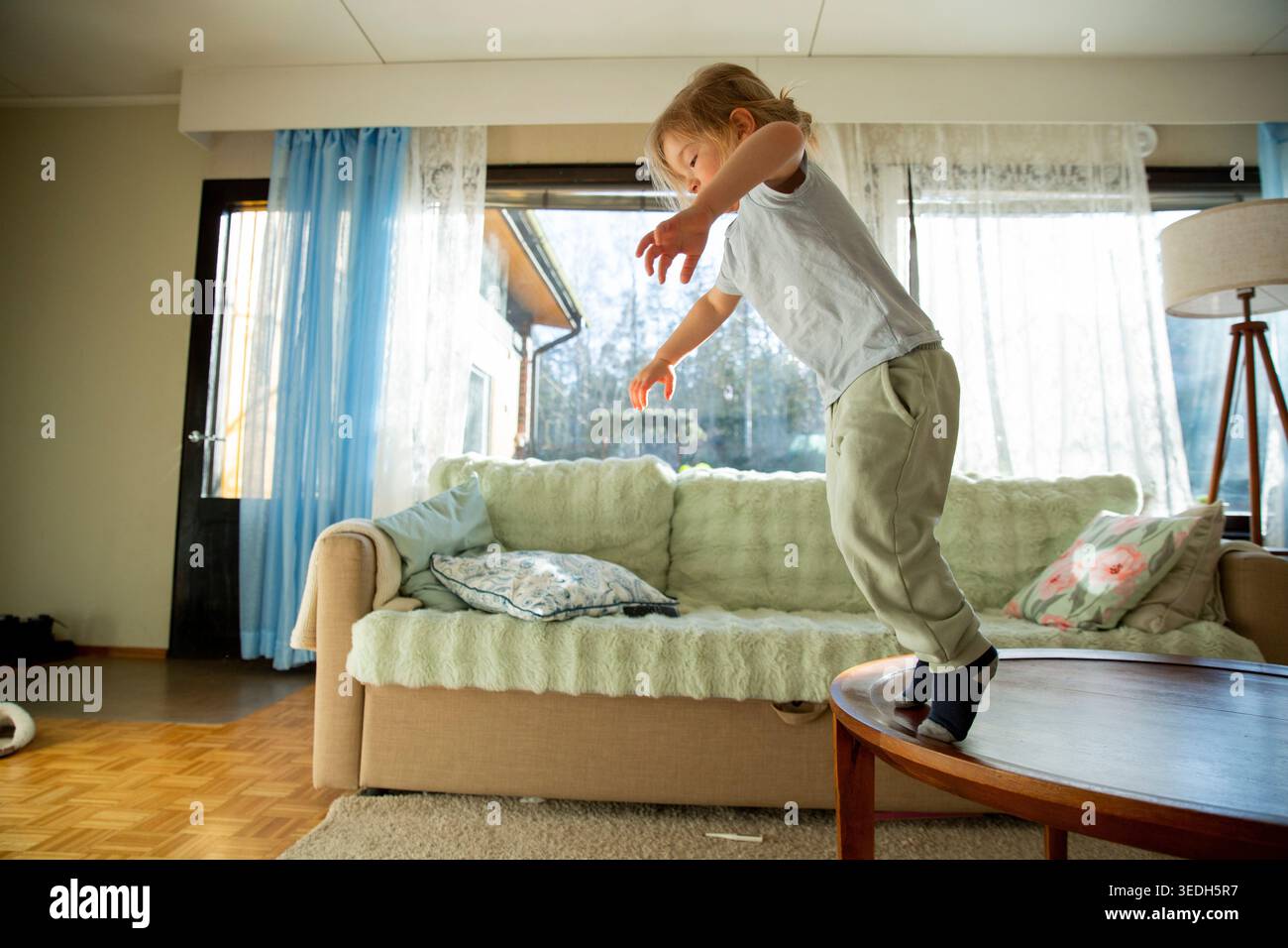 Niedrigwinkelaufnahme eines fröhlichen kleinen Kindes mit langen Haaren, das vom Sofa springt, voller Energie und verspieltem Lachen während eines hellen morgens zu Hause Stockfoto
