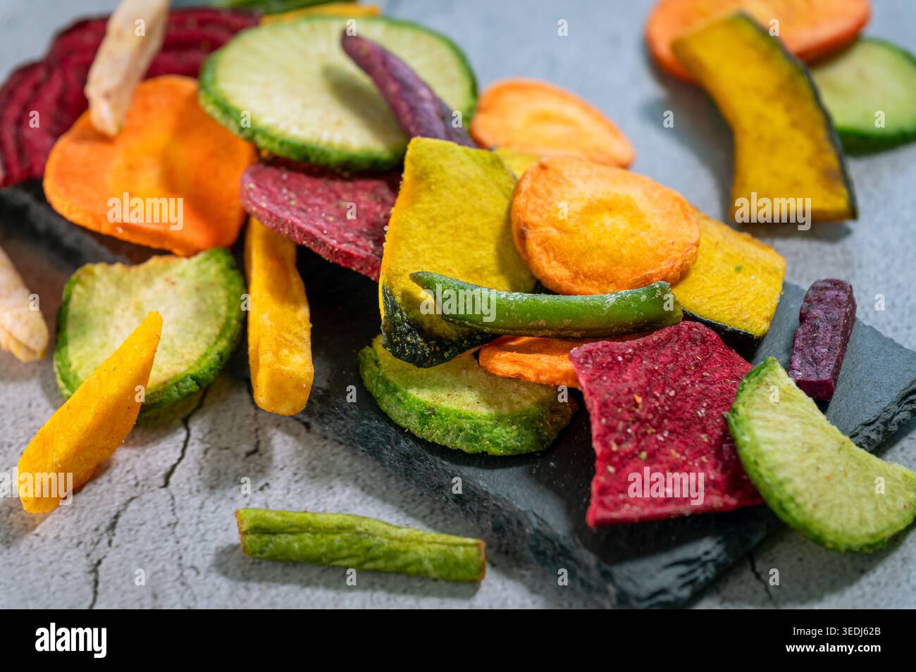 Auswahl an bunten getrockneten Gemüsechips, darunter Rote Bete, Karotten, Zucchini und Süßkartoffeln, die auf einer Schiefer- und Steinoberfläche verstreut sind. Stockfoto