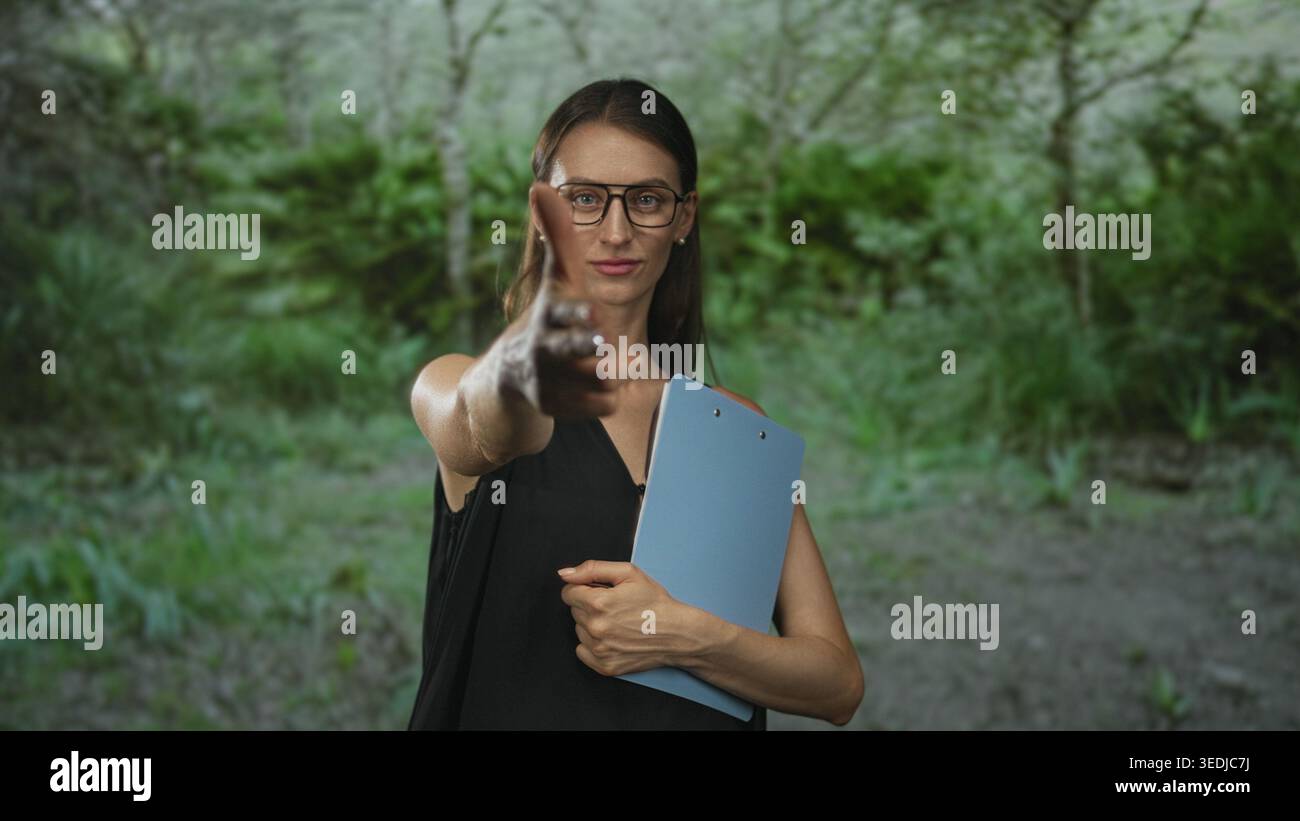 Frau, die eine Brille trägt, mit den Händen blaues Klemmbrett hält, zeigt den Finger nach vorne im Wald; Selbstvertrauen. Stockfoto