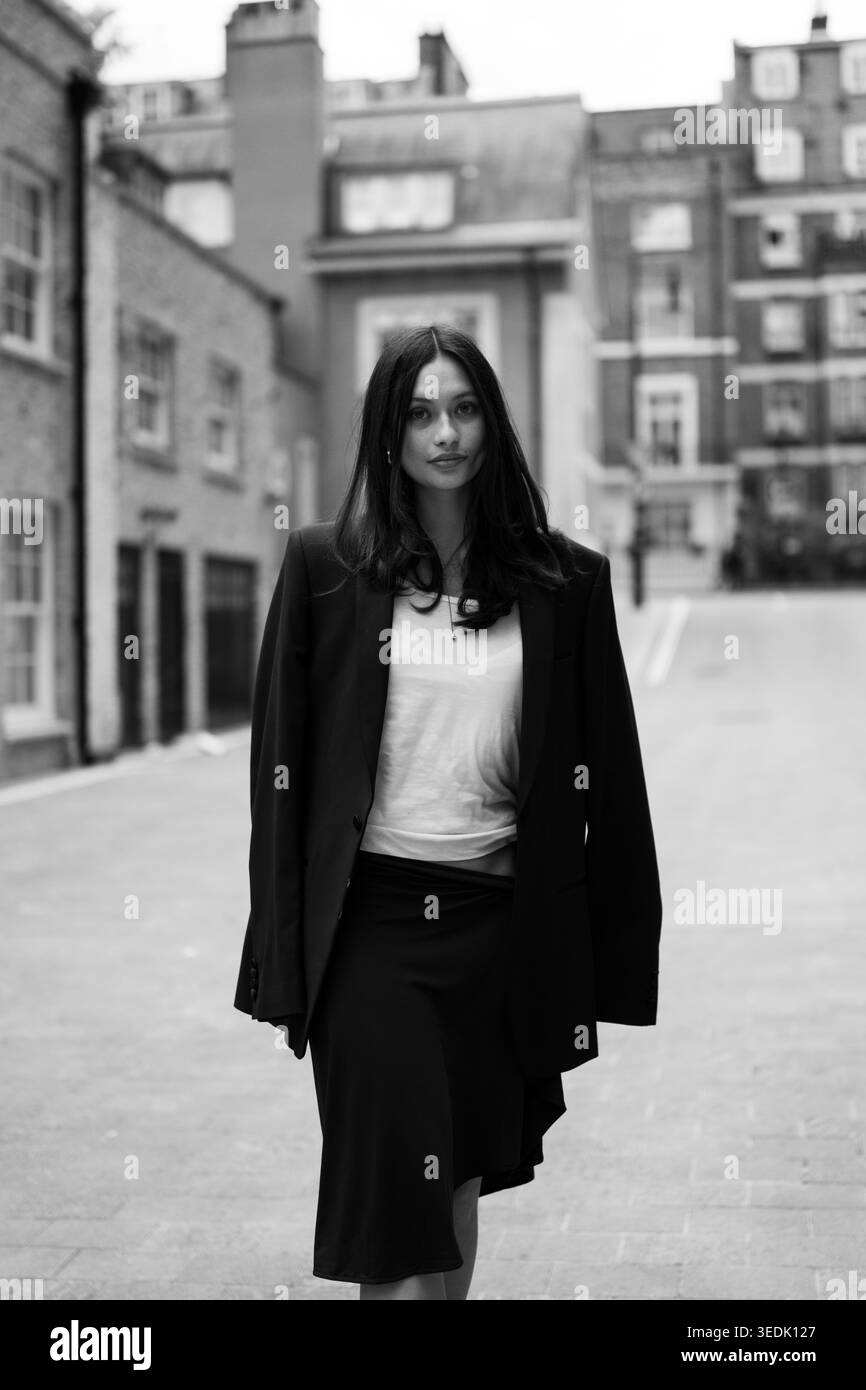 Junge Frau in schwarzem Blazer und weißem Oberteil, die draußen vor einer Ziegelwand in urbaner Umgebung posiert, modernes Modeporträt in natürlichem Licht. Stockfoto