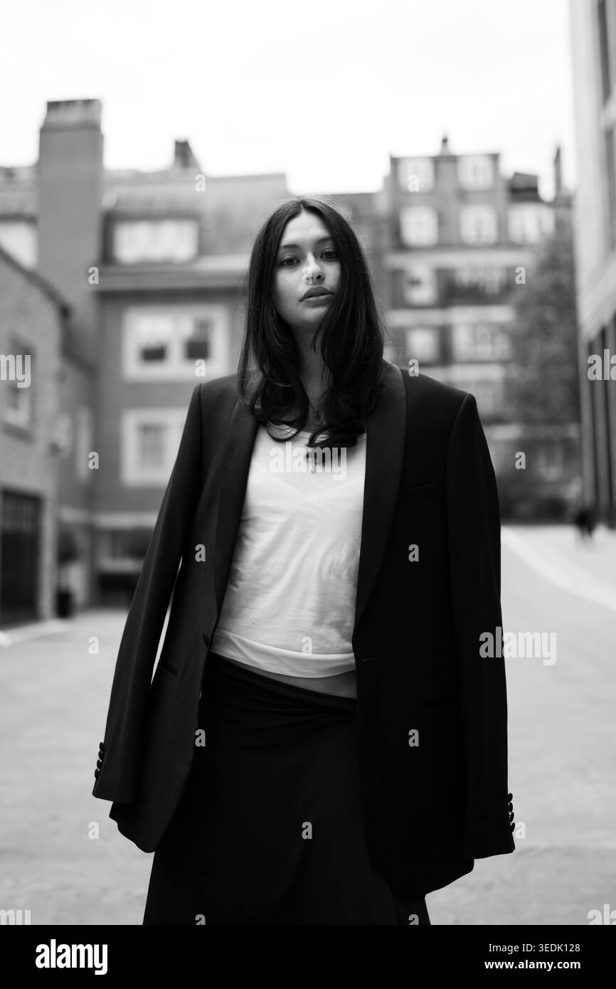 Junge Frau in schwarzem Blazer und weißem Oberteil, die draußen vor einer Ziegelwand in urbaner Umgebung posiert, modernes Modeporträt in natürlichem Licht. Stockfoto
