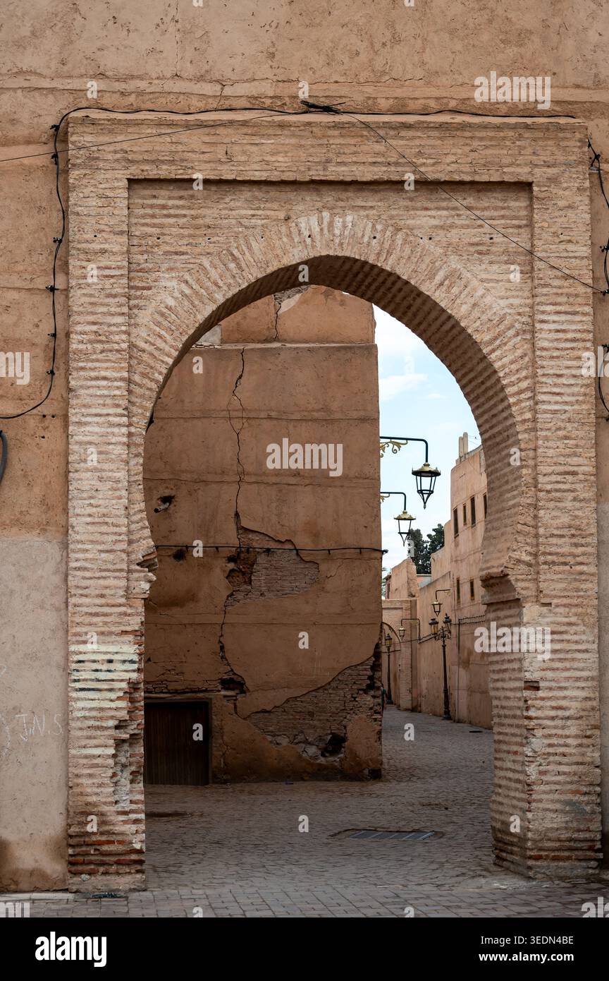 Sonnendurchfluteter Bogen in der Medina von Marrakesch umrahmt eine enge Kopfsteinpflasterstraße mit verwitterten Lehmziegelwänden, traditionellen Laternen und einem Sinn für Geschichte, dezember Stockfoto