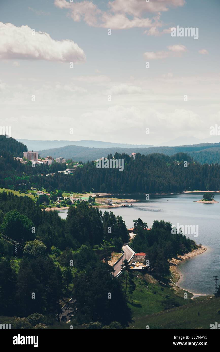 Malerische Seenlandschaft mit bewaldeten Hügeln, einem kleinen Dorf und einer gewundenen Straße, aus der Vogelperspektive auf die friedliche Landschaft mit Wasser und Bergen bei sanften Tagen Stockfoto