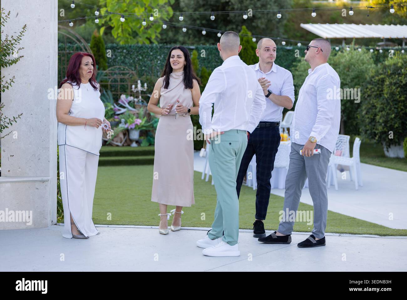Gotse Delchev, Bulgarien - 07 08 2025: Gruppe von Erwachsenen, die sich bei einer Gartenveranstaltung im Freien mit dekorativen Lichtern und Grün unterhalten. Freizeit g Stockfoto