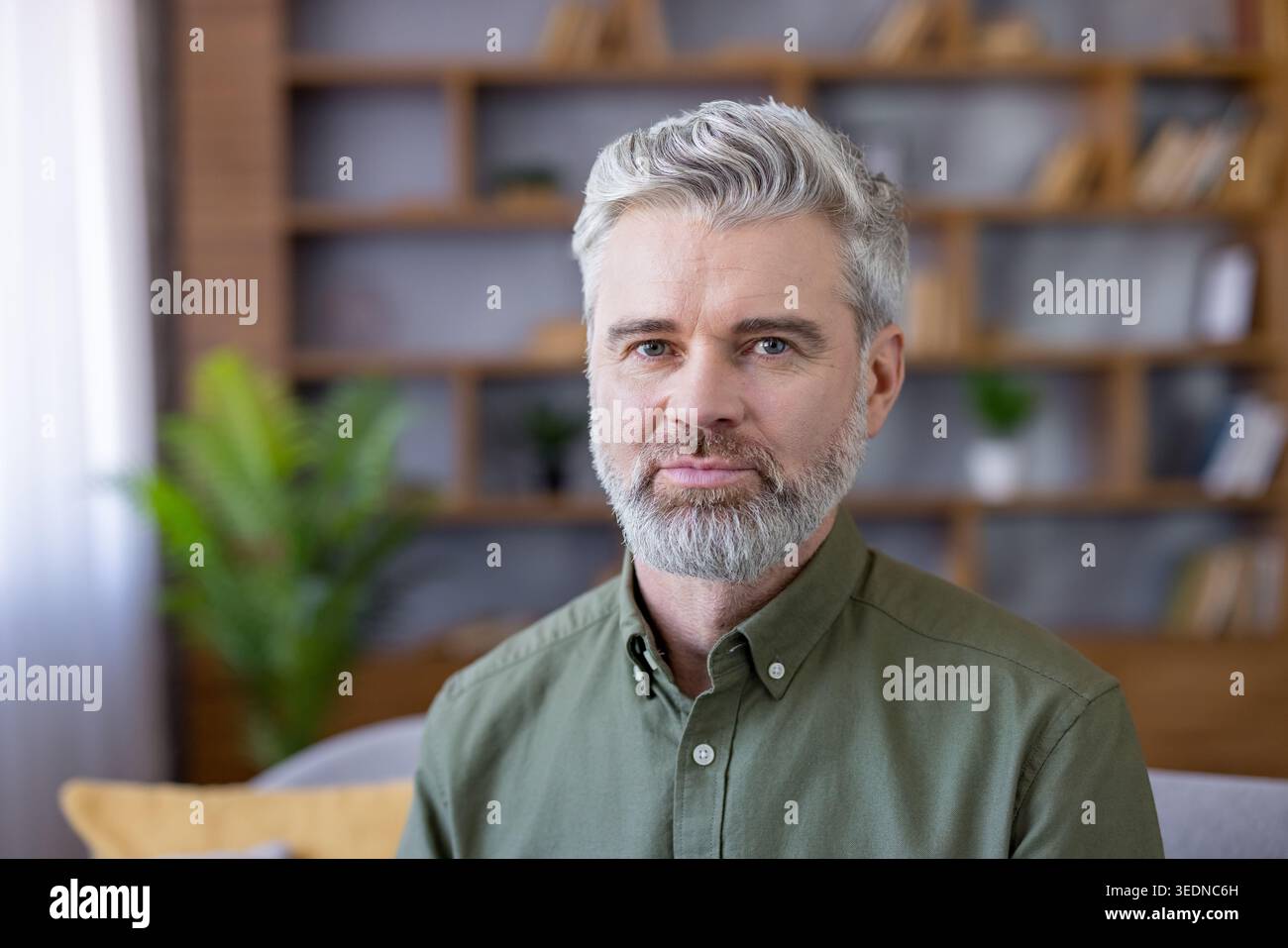Selbstbewusster Mann mittleren Alters mit grauen Haaren und Bart, der vor verschwommenem Hintergrund im Wohnzimmer oder Home Office sitzt und Professionalität, Erfahrung und Fernarbeit zeigt Stockfoto