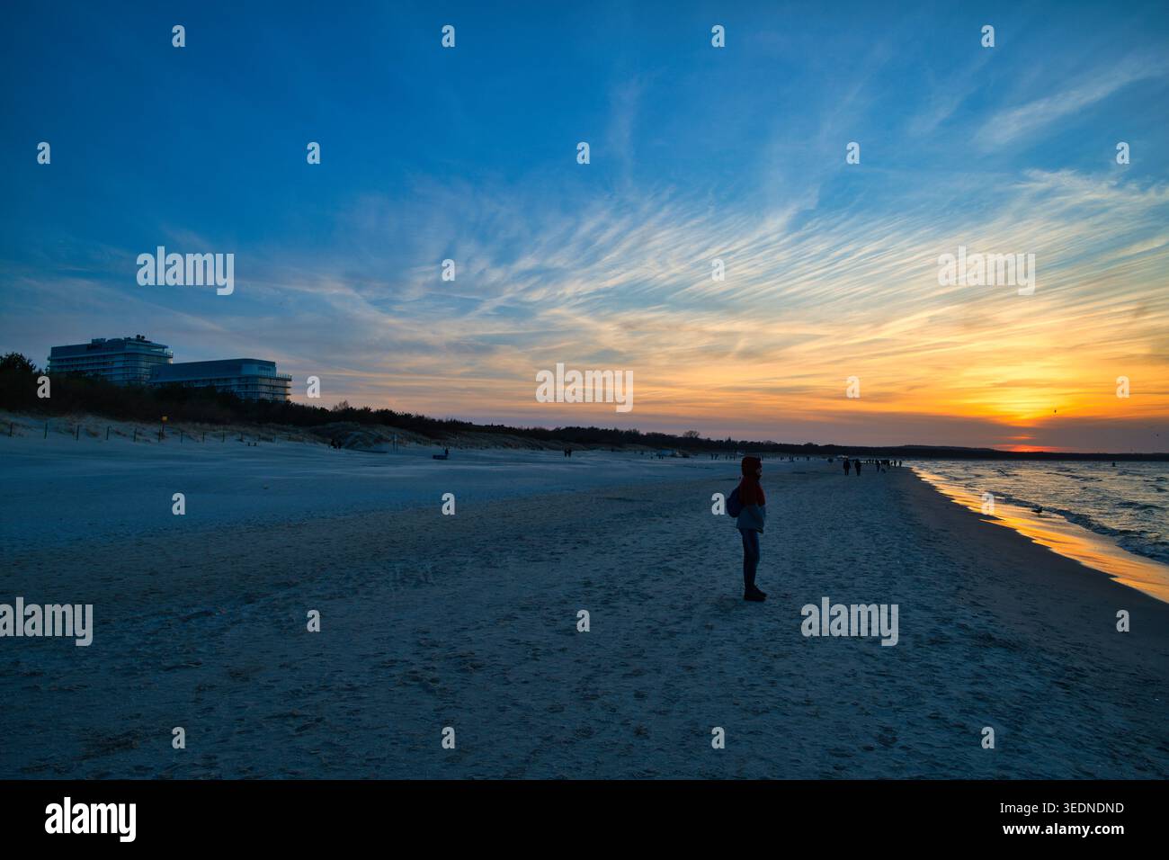 Einsame Figur, die den farbenfrohen Sonnenuntergang über Sandstrand und die ruhige Küste des Ozeans mit weit entfernten Gebäuden beobachtet - die Küstenstadt Świnoujście. Stockfoto