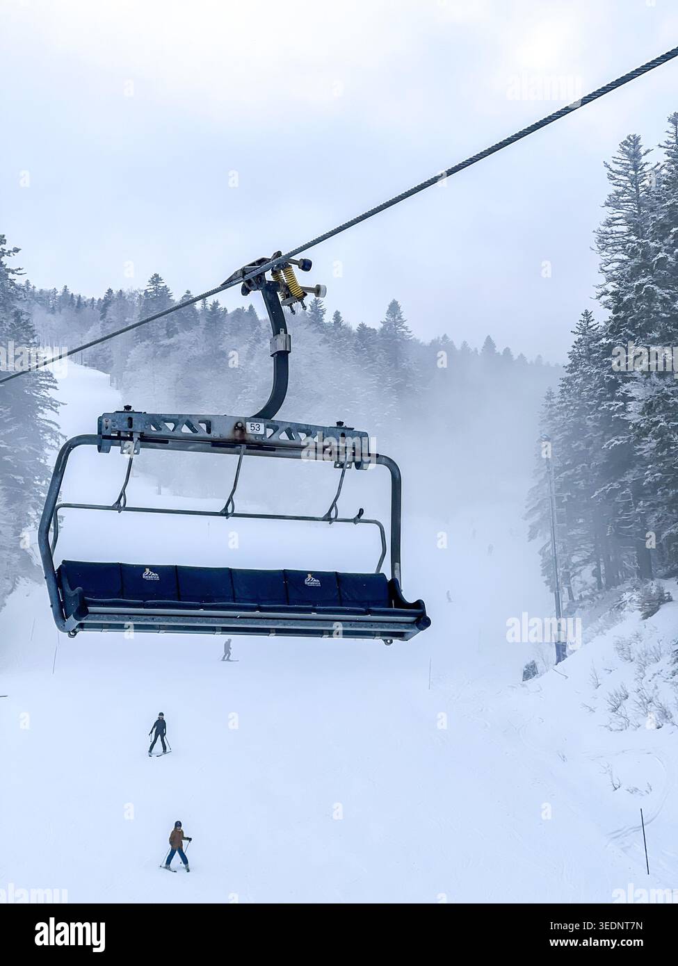 Leerer Sessellift über schneebedeckten Pisten mit Skifahrern unten im nebeligen Bjelašnica bei Sarajevo, Bosnien und Herzegowina. - Smartphone-aufgenommenes Stockfoto