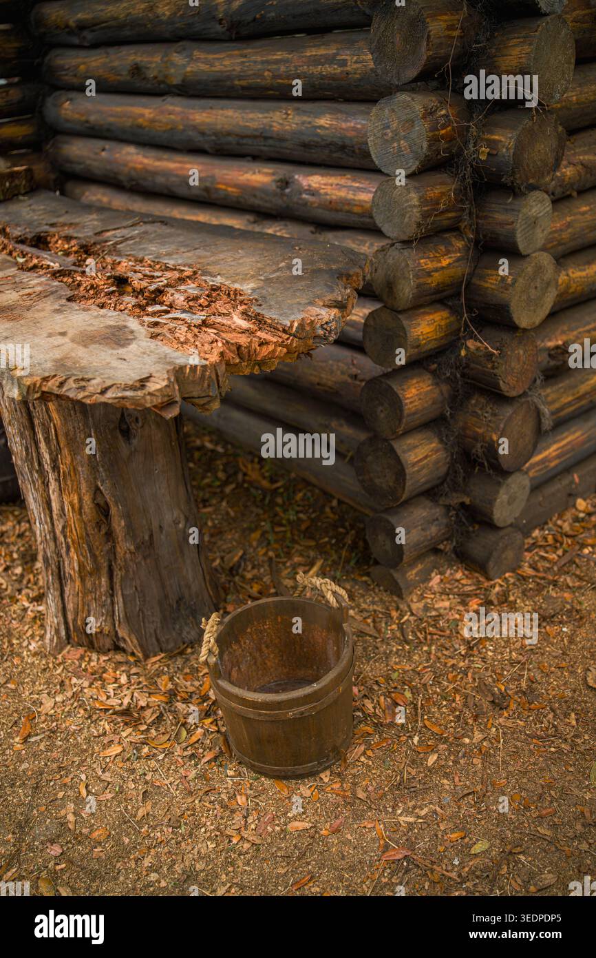 Eine geschnitzte Tischplatte aus rohem Holz neben einer traditionellen Blockhüttenwand mit einem antiken rostigen Eimer, der auf trocken gefallenen Eichenblättern ruht. Stockfoto