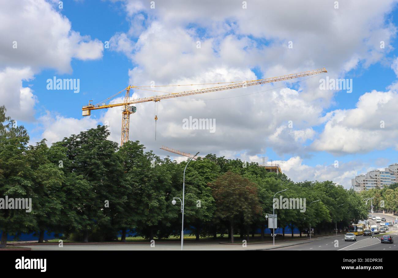 Der Himmel hält Wolken, was auf einen arbeitsreichen Tag hindeutet. Bäume säumen die Straße unter Baumaschinen. Weit entfernte Fahrzeuge bewegen sich langsam durch den städtischen Raum Stockfoto