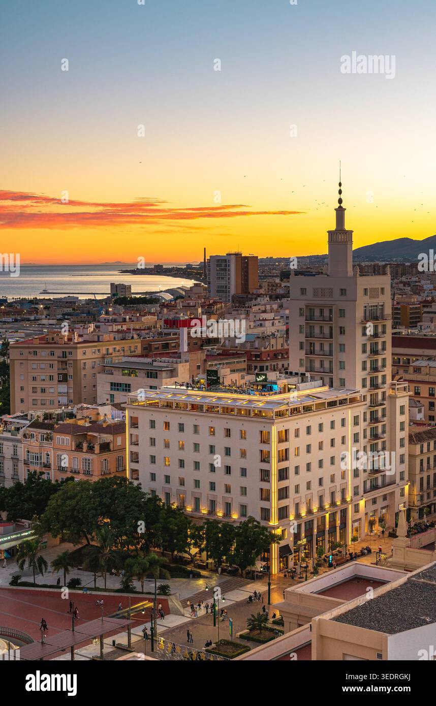 Blick auf Malaga, Spanien bei Sonnenuntergang, mit beleuchteten mittelhohen Gebäuden, einem markanten Turm, einer Dachterrasse und einem Stadtplatz mit Baum Stockfoto