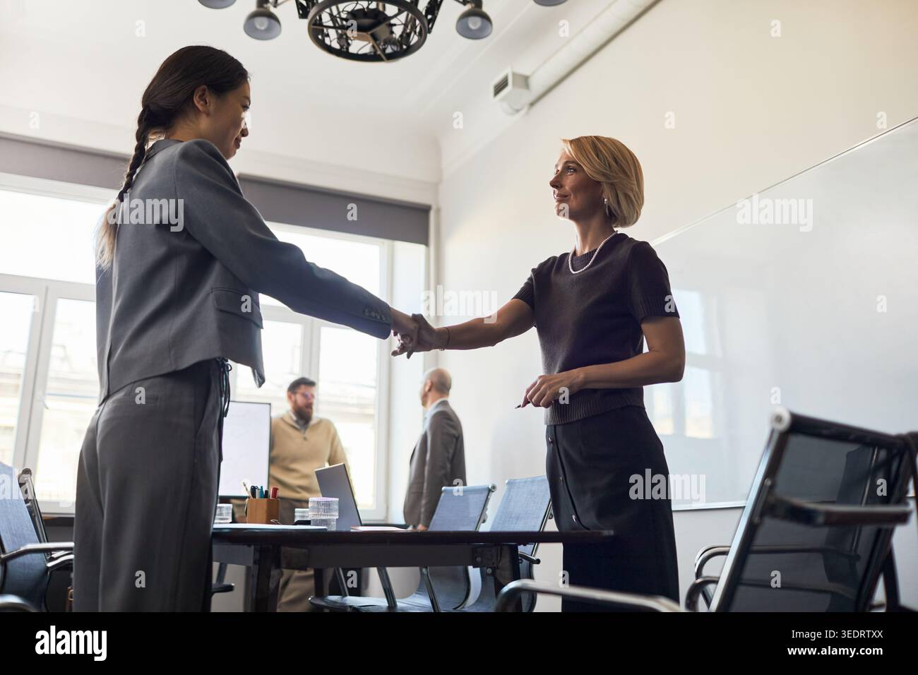 Weitwinkelaufnahme von zwei Geschäftsfrauen, die sich bei einem Büromeeting die Hand schütteln, um Partnerschaft und Verhandlungen zu signalisieren. Ideal für geschäftliche Teamarbeit oder Einstellungen Stockfoto