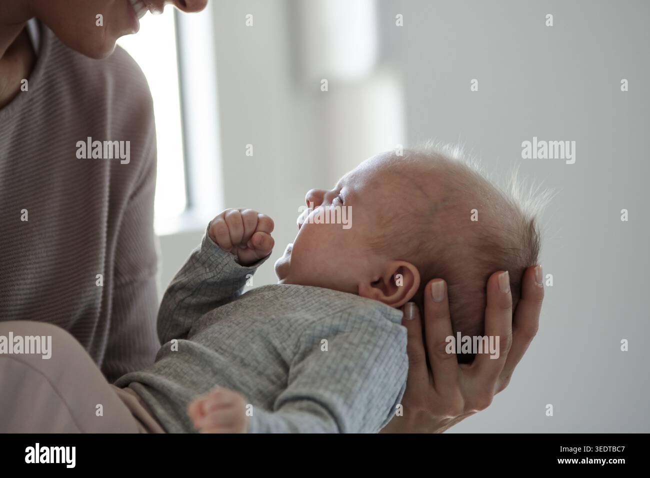 Eine Mutter hält ihr Baby sanft in einem warmen Zimmer. Das Baby schaut zu ihr auf, während es kleine Gesten mit seinen Händen macht. Stockfoto