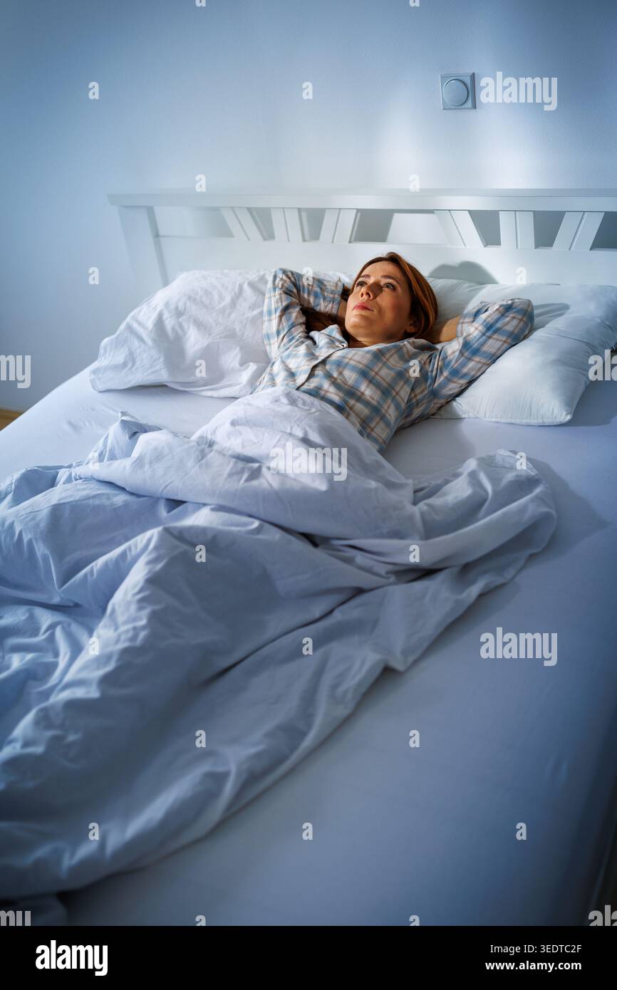 Eine Frau liegt im Bett mit weißen Laken und Kissen. Sie sieht in Gedanken verloren aus und starrt an die Decke. Das Morgenlicht erfüllt den Raum. Stockfoto