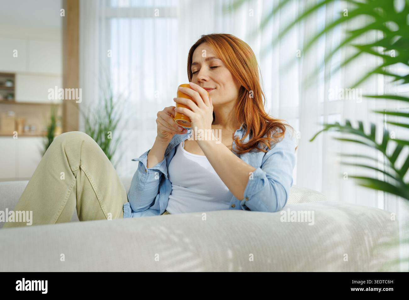Eine Frau sitzt auf einer Couch, hält ein warmes Getränk und lächelt, während sie sich tagsüber in ihrem Haus entspannt. Sonnenlicht erfüllt den Raum. Stockfoto
