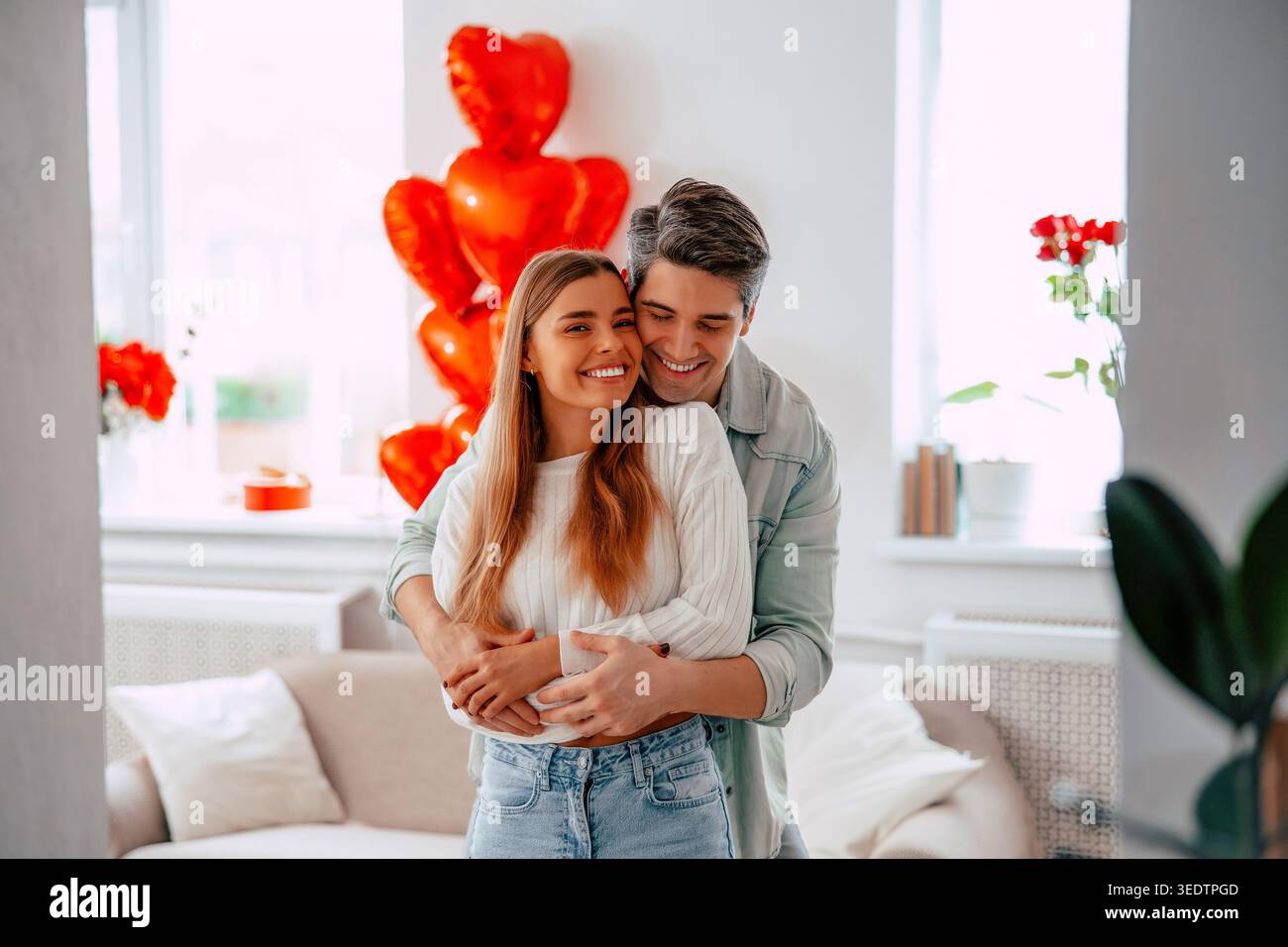 Glückliches junges Paar, das sich zu Hause umarmt, romantische Partner, die sich mit roten Herzballons in einem gemütlichen, modernen Wohnzimmer einen zärtlichen Moment teilen Stockfoto