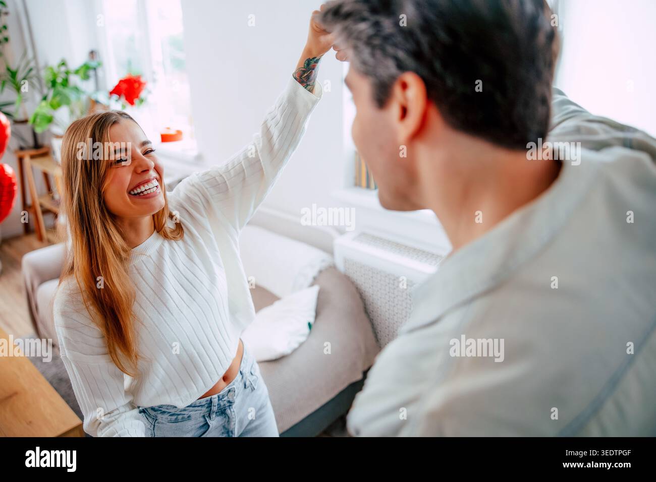 Glückliche junge Frau lächelt beim Tanzen mit Freund zu Hause, romantisches Paar genießt spielerische Momente in hellem, modernem Wohnzimmer Stockfoto