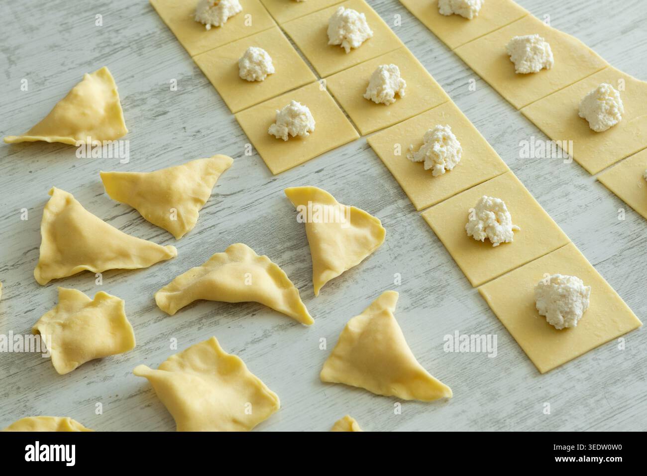 Pasta-Teig-Quadrate mit Weißkäsefüllung und gefalteten Ravioli auf Holzoberfläche. Stockfoto