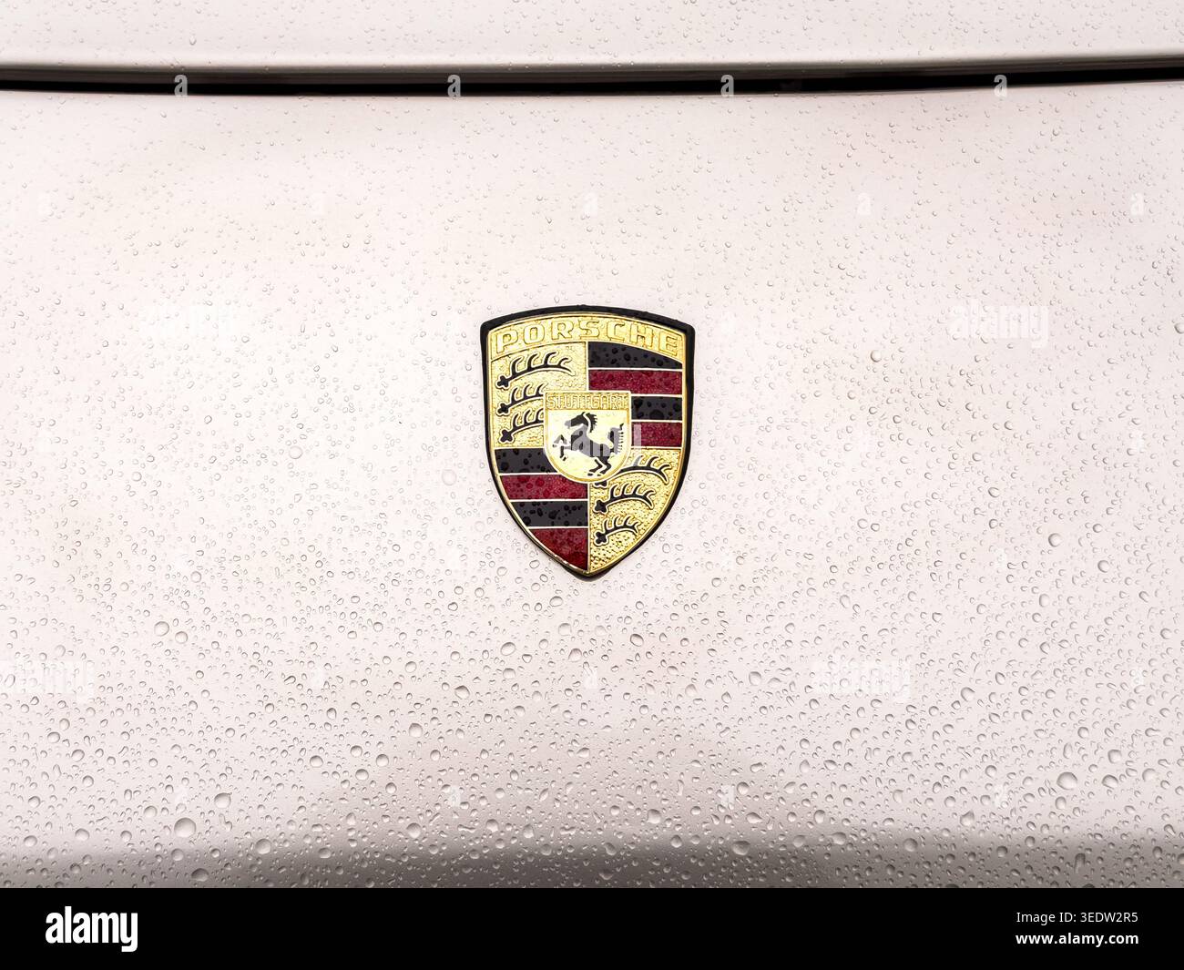 Nahaufnahme des ikonischen goldenen und roten Porsche-Wappens auf einer silbernen, metallischen Autohaube, die nach einem Sturm mit frischen Regentropfen bedeckt ist Stockfoto