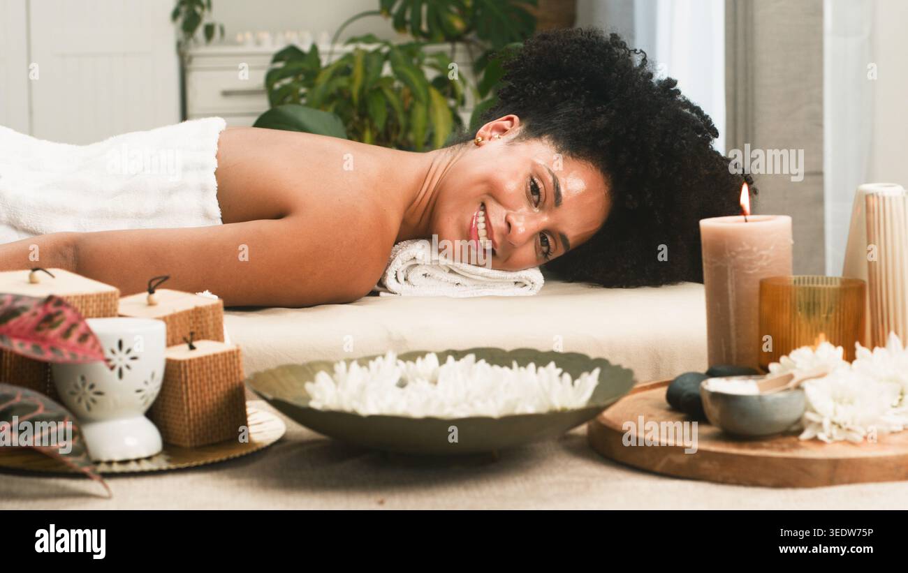 Entspannende Spa-Massage mit lächelnder Frau, umgeben von Kerzen, Blumen und Spa-Zubehör Stockfoto