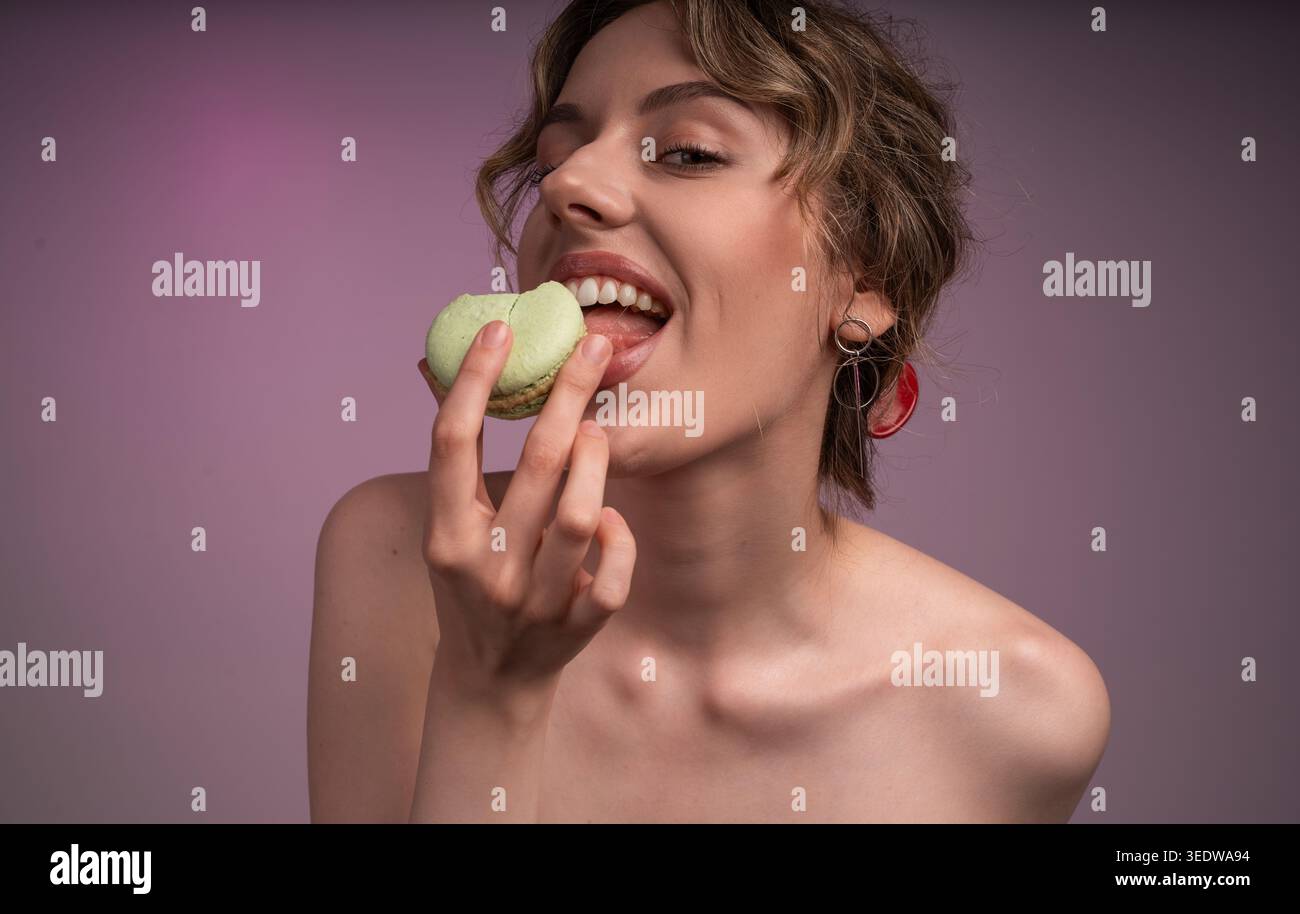 Junge Frau genießt süßen grünen Macaron, schmeckt Dessert mit fröhlichem Ausdruck und offenem Mund Stockfoto