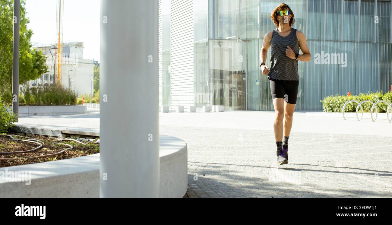 Ein Mann läuft nachmittags auf einem Weg neben einem modernen Gebäude. Er trägt Laufshorts und ein Tanktop. Die Sonne reflektiert von der Glasstruktur Stockfoto
