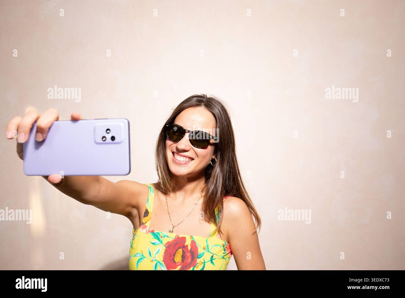 Frau lächelt, hält ein Smartphone und macht ein Selfie, während sie eine Sonnenbrille und ein Blumenkleid trägt Stockfoto