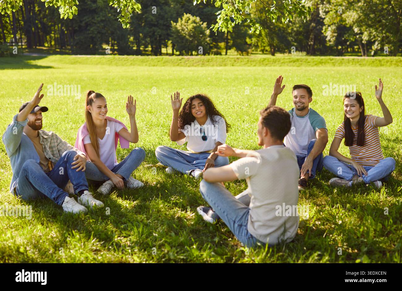 Gruppe junger multiethnischer Erwachsener, die im Kreis auf dem Gras sitzen, die Hände heben, sich beteiligen Stockfoto