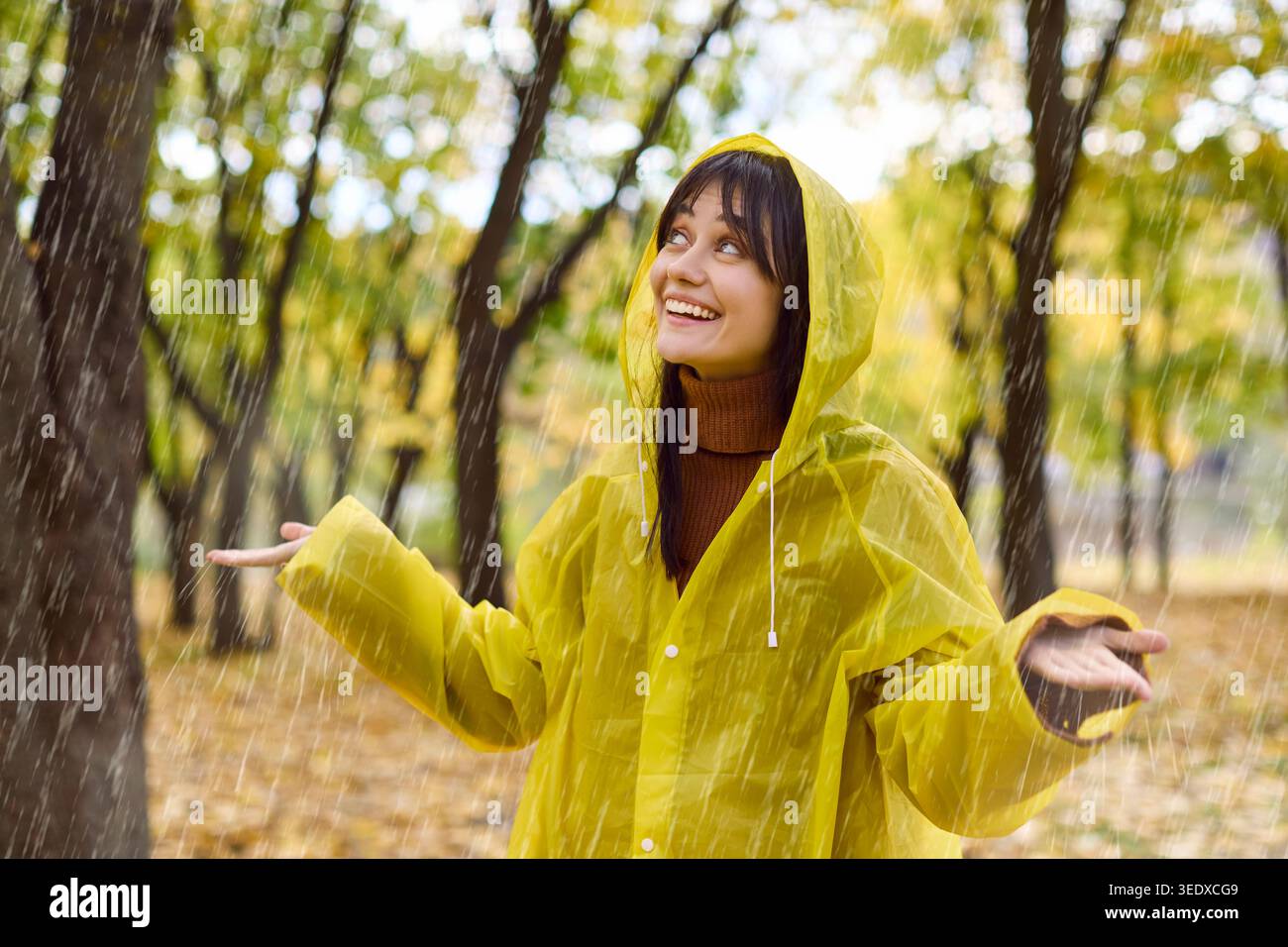 Glückliche junge Frau in gelbem Regenmantel genießt Herbstregen im grünen Park draußen Stockfoto