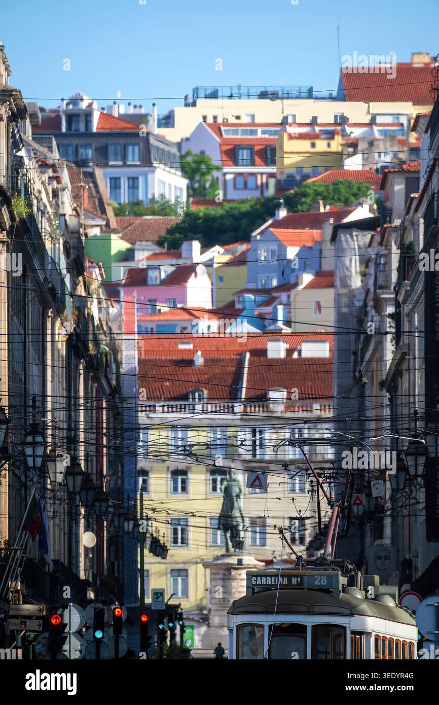 Blick auf eine traditionelle gelbe Straßenbahn auf einer schmalen Straße, die zur Praca do Comercio mit farbenfrohen Häusern auf dem Hügel in Lissabon, Lissabon, Portugal führt. Stockfoto