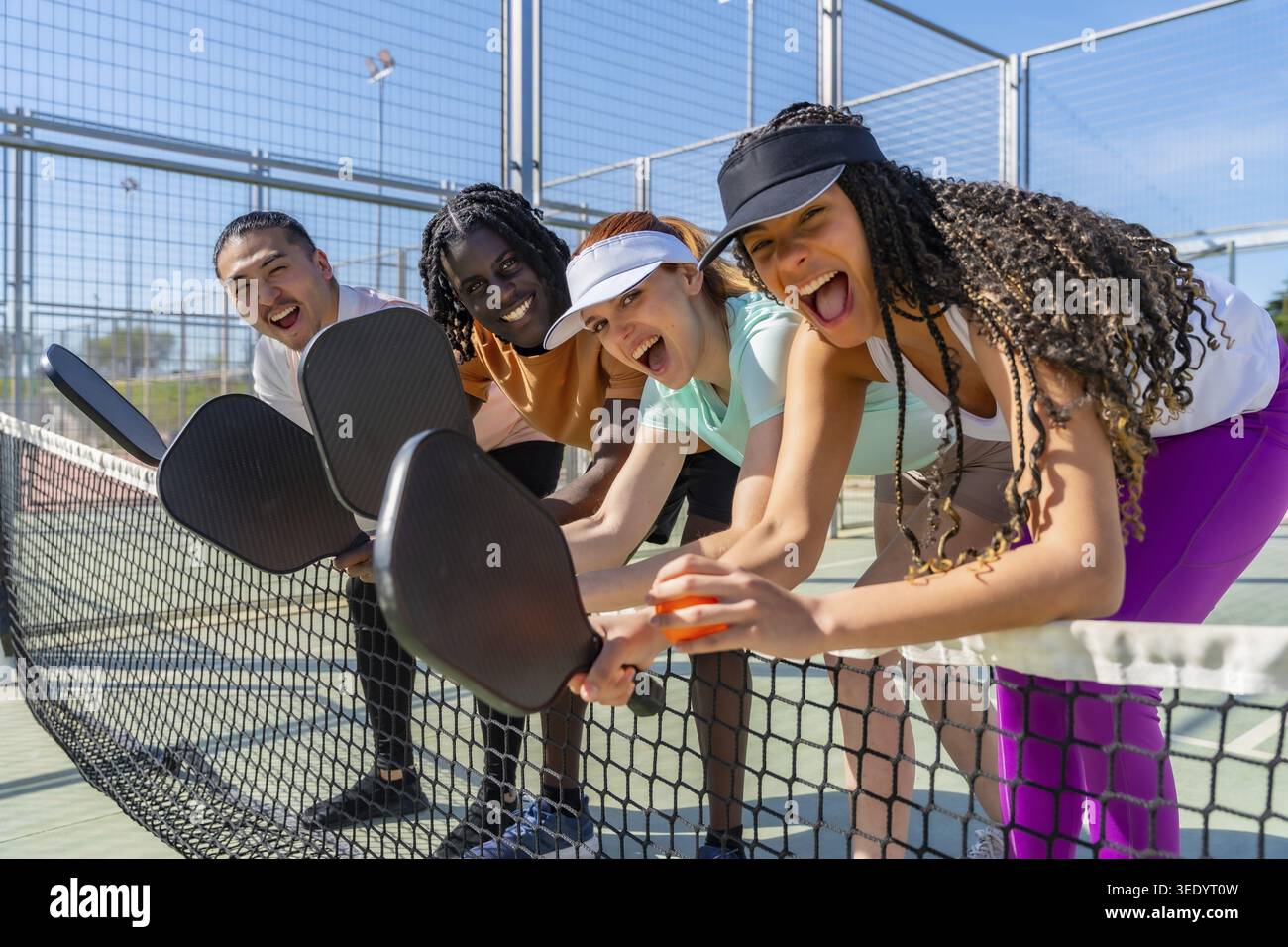 Verschiedene Freunde lehnen sich spielerisch über ein Pickleball-Netz, lachen und halten Paddel und genießen gemeinsam ein aufregendes und unterhaltsames Outdoor-Sportspiel auf einem Stockfoto