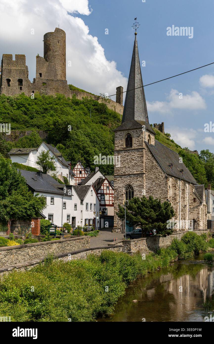 Blick auf die historische Steinkirche und Fachwerkhäuser entlang des Flusses mit mittelalterlichen Burgruinen auf einem üppig grünen Hügel in Monreal, Rheinland-Palat Stockfoto