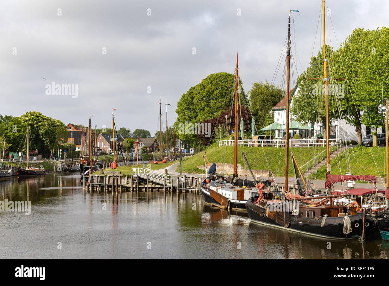 Blick auf den Hafen Harlesiel mit traditionellen Segelbooten entlang des Kanals, umgeben von üppigen grünen Bäumen und historischen Gebäuden in Wittmund, Lowe Stockfoto