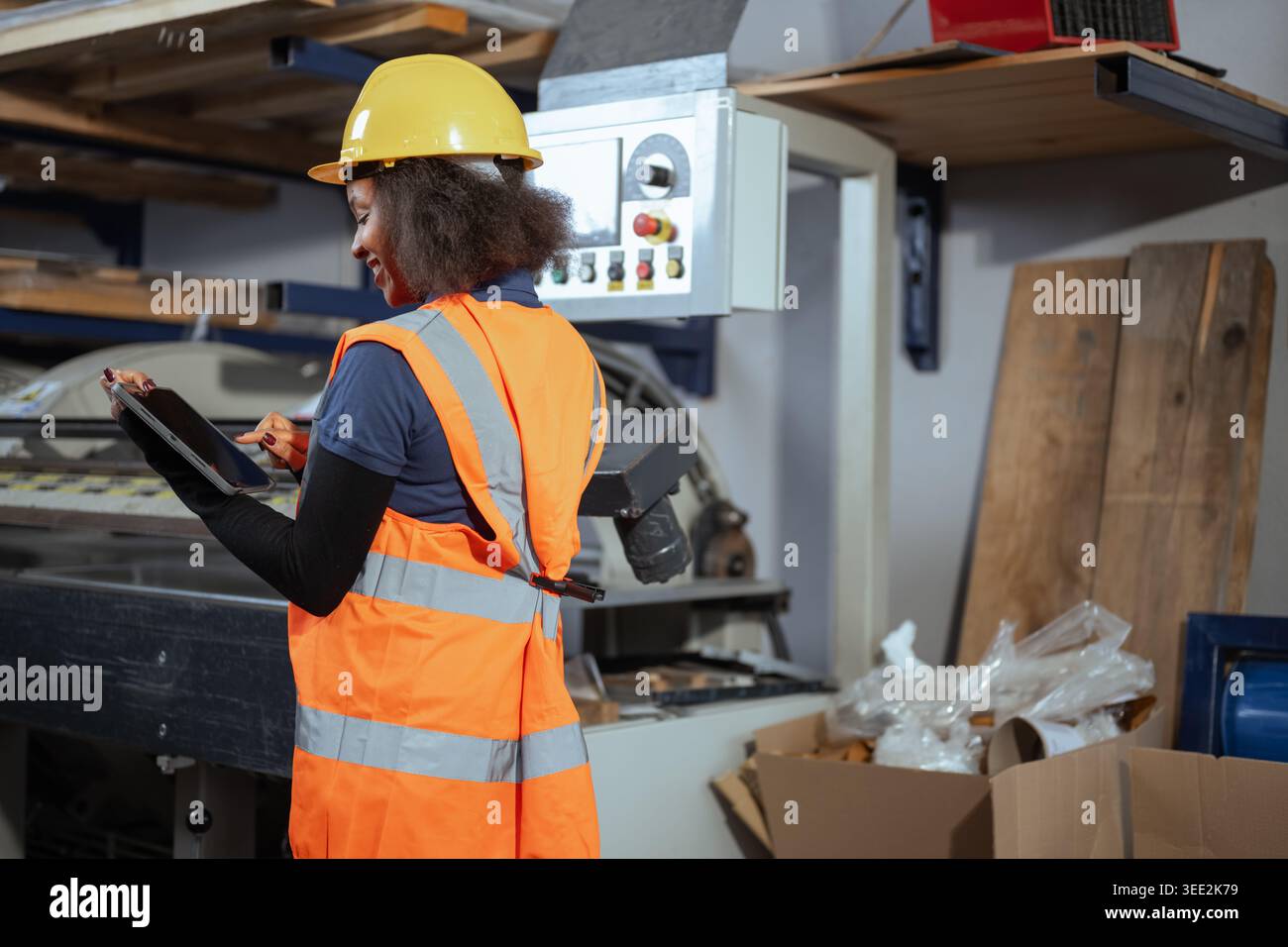 Eine afroamerikanische Frau mit gelbem Schutzhelm benutzt ein digitales Tablet, während sie Maschinen in einer Werkseinstellung überprüft. Stockfoto