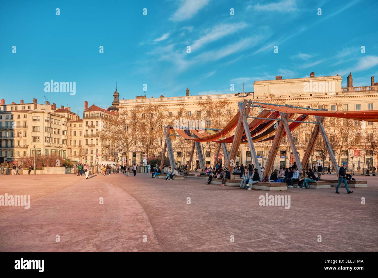 Lyon, Frankreich - 19. März 2026: Menschen, die sich bei Sonnenuntergang unter modernen Holzschattierungen am Place Bellecour im historischen Stadtzentrum entspannen Stockfoto