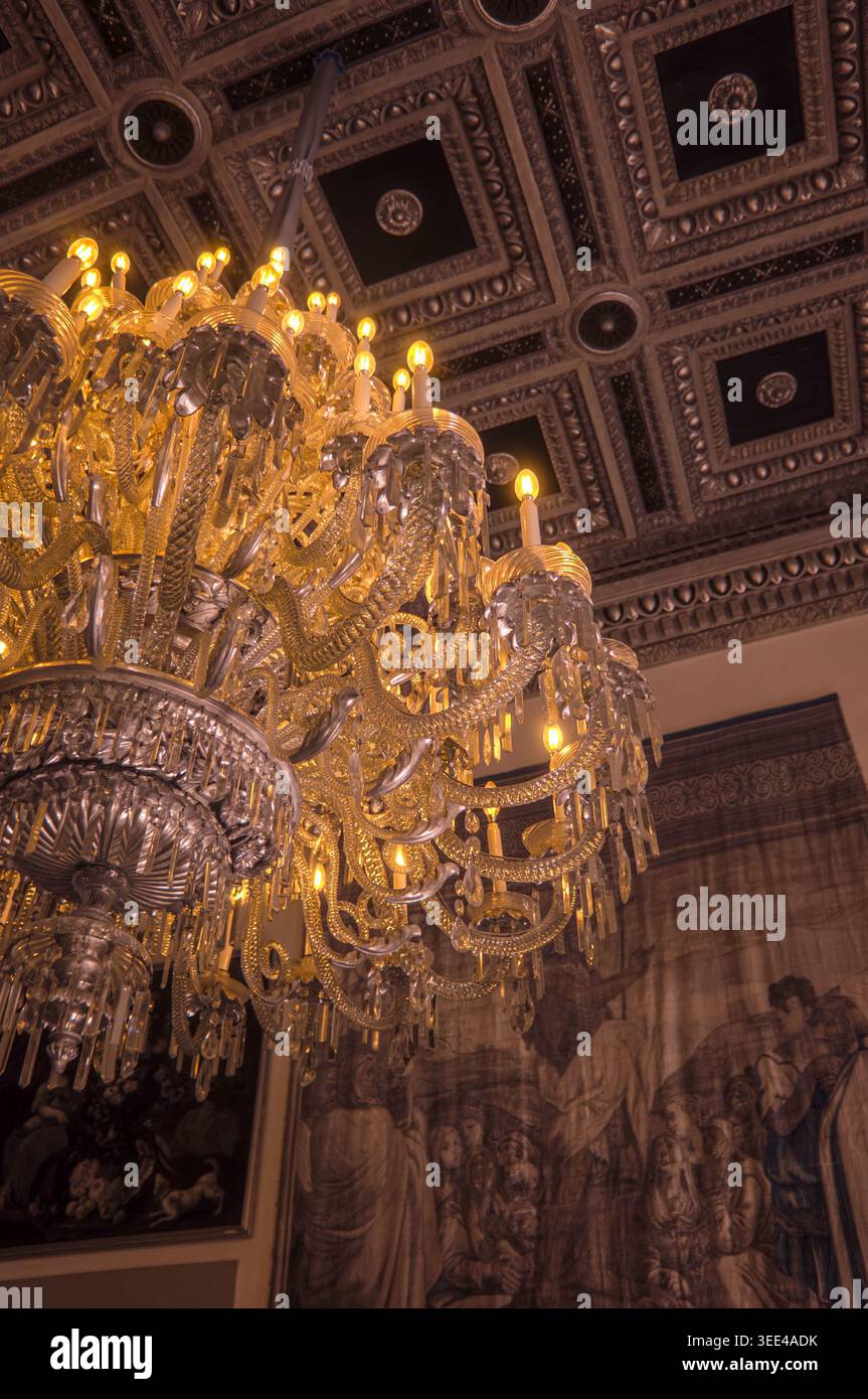 Ein Blick nach oben auf einen großen, raffinierten Kronleuchter mit komplizierten Gold- und Kristalldetails, die einen warmen, goldenen Glanz ausstrahlen. Der Kronleuchter hängt von einem dar Stockfoto