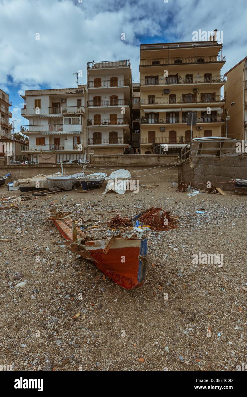 Aus nächster Nähe sehen Sie ein abgenutztes, rot-blaues Fischerboot, das auf einem mit Trümmern übersäten, düsteren Strand unter einem bewölkten Himmel ruht. Mehrere mehrstöckige Wohngebäude Stockfoto