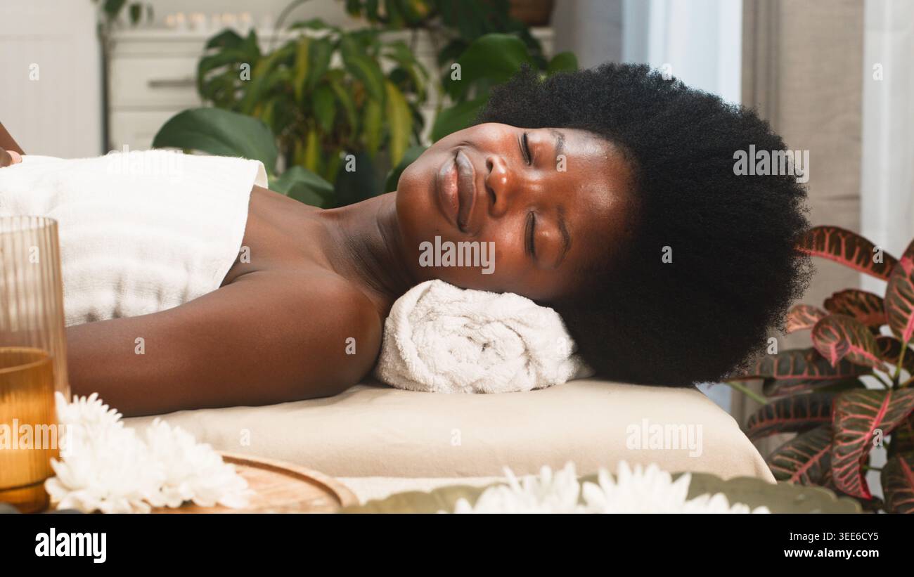 Entspannte Frau, die eine Spa-Behandlung mit natürlichem Haar auf gerolltem Handtuch in ruhiger Umgebung erhält Stockfoto