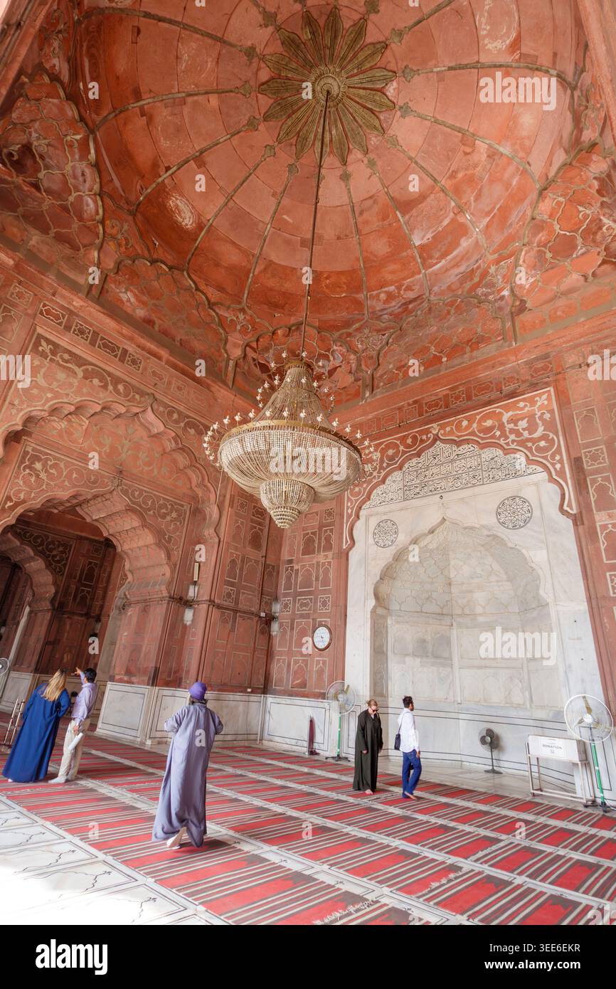 Innenraum der großen Jama Masjid Moschee in Delhi Indien mit roten Sandsteinbögen und einem großen zentralen Kronleuchter mit Besuchern im Gebetssaal Stockfoto