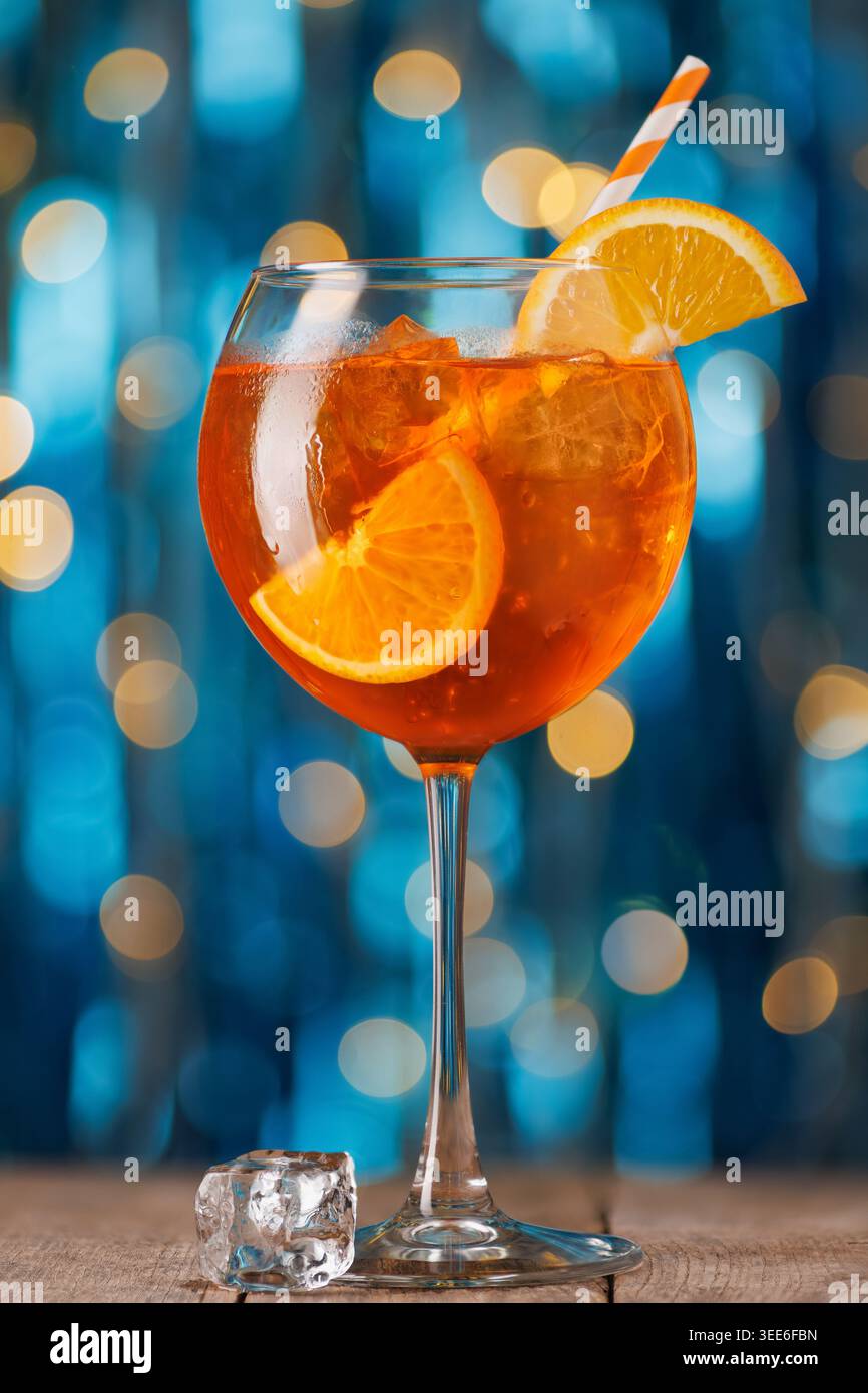 Aperol spritzt Cocktail mit Orangenscheibe und Eis auf rustikalem Holztisch, festliche blaue und goldene Bokeh-Lichter Stockfoto