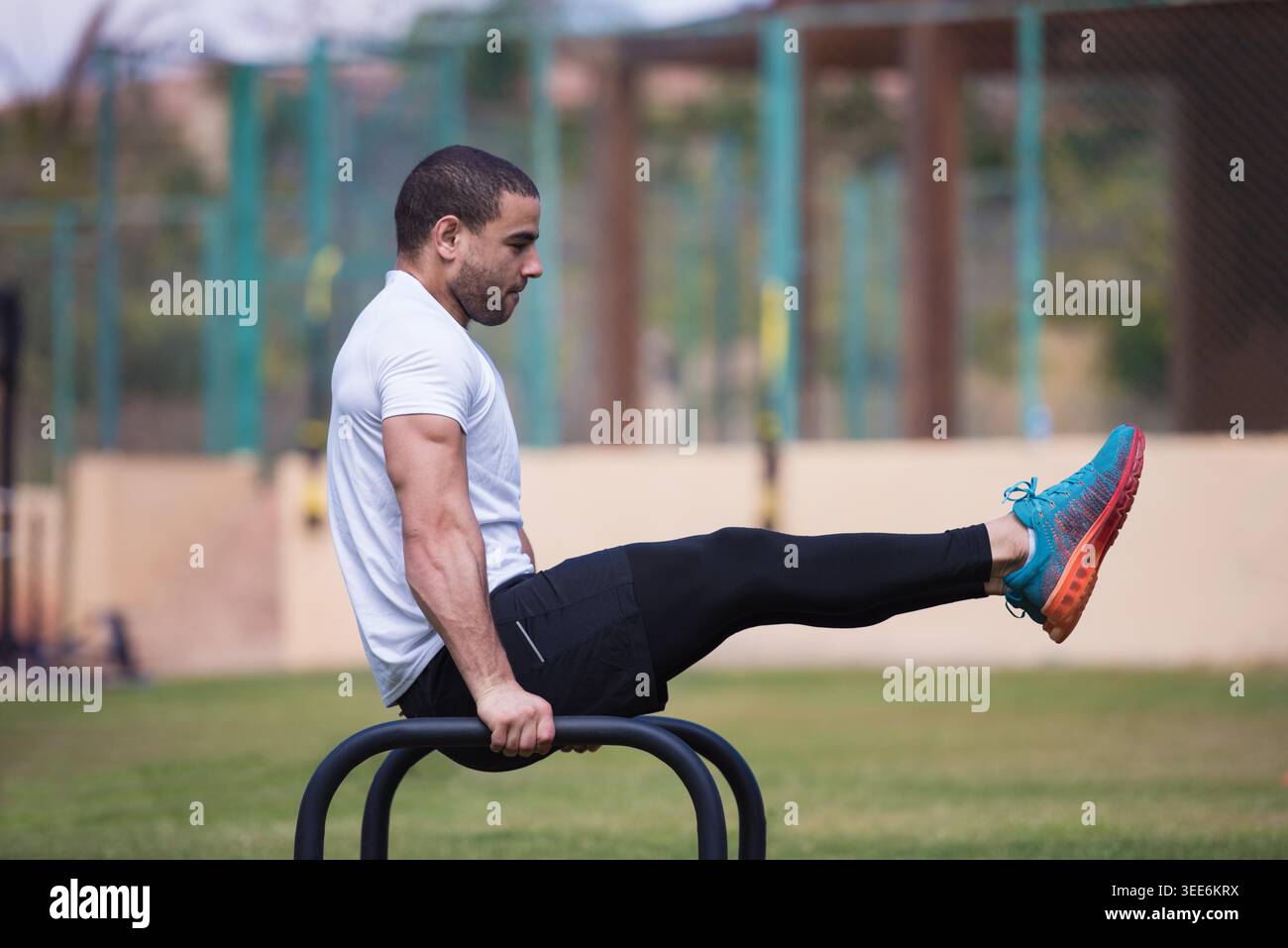 Sportlicher Mann beim L-Sit auf parallelen Bars in einem Fitnessstudio im Freien, konzentriert sich auf Kraft, Calisthenics und Crossfit-Training mit einem Freund, zeigt Balance, Core Control und disziplinierte Workout-Form. Stockfoto