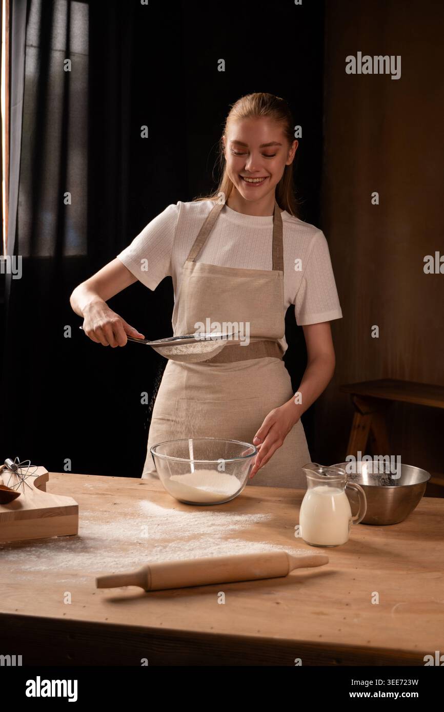Lächelnde Frau siebt Mehl in eine Schüssel auf einem Holztisch und bereitet Zutaten zum Backen vor Stockfoto
