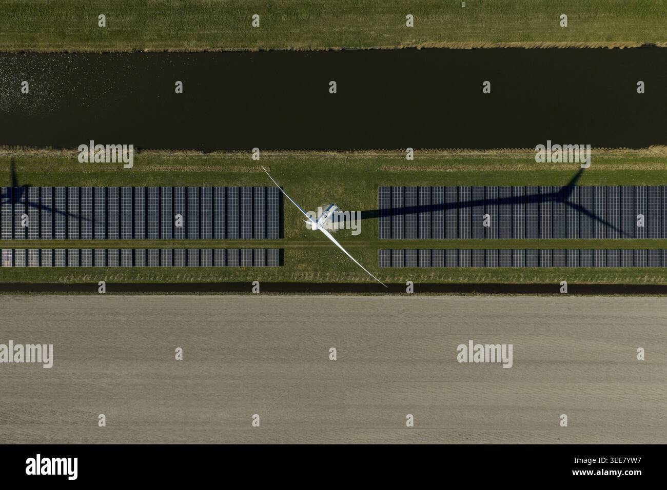 Luftaufnahme einer Windkraftanlage, die einen langen Schatten über Reihen von Solarpaneelen neben einem Kanal und landwirtschaftlichen Feldern in den Niederlanden wirft. Stockfoto