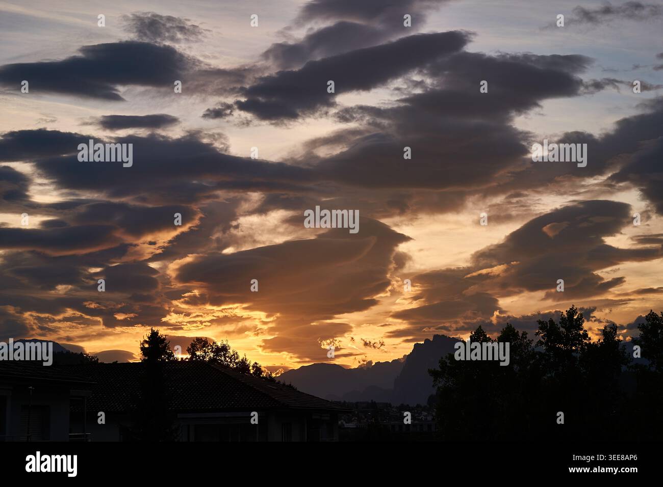 Der dramatische orangene Sonnenuntergang beleuchtet Lentikularwolken über Silhouettendächern, Bäumen und weit entfernten Berggipfeln. Stockfoto
