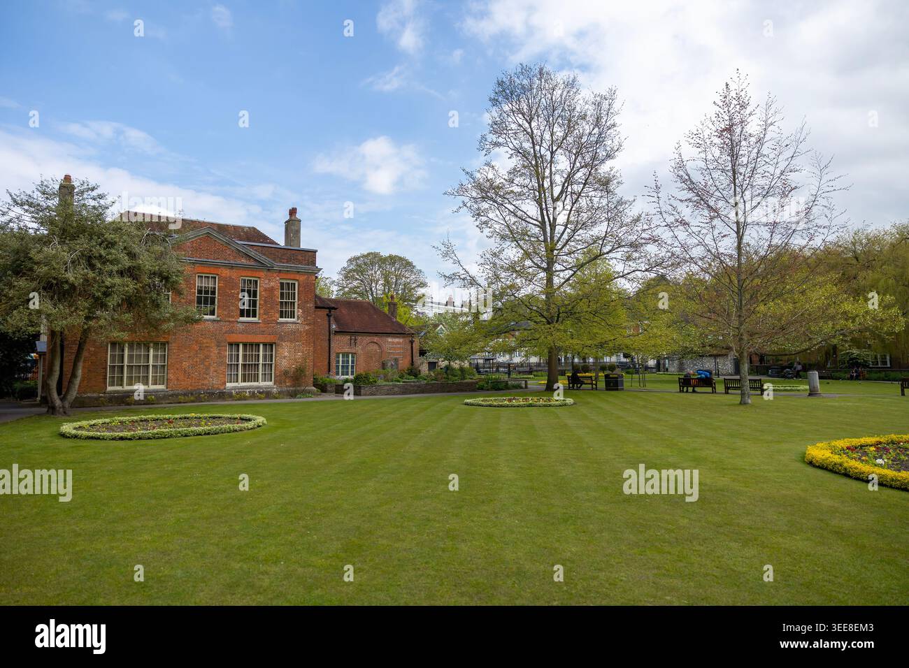 Rotes Backsteingebäude und Landschaftsgärten in Winchester, Hampshire, England – Blick auf den Frühling mit Rasen, Bäumen und Bänken. Stockfoto