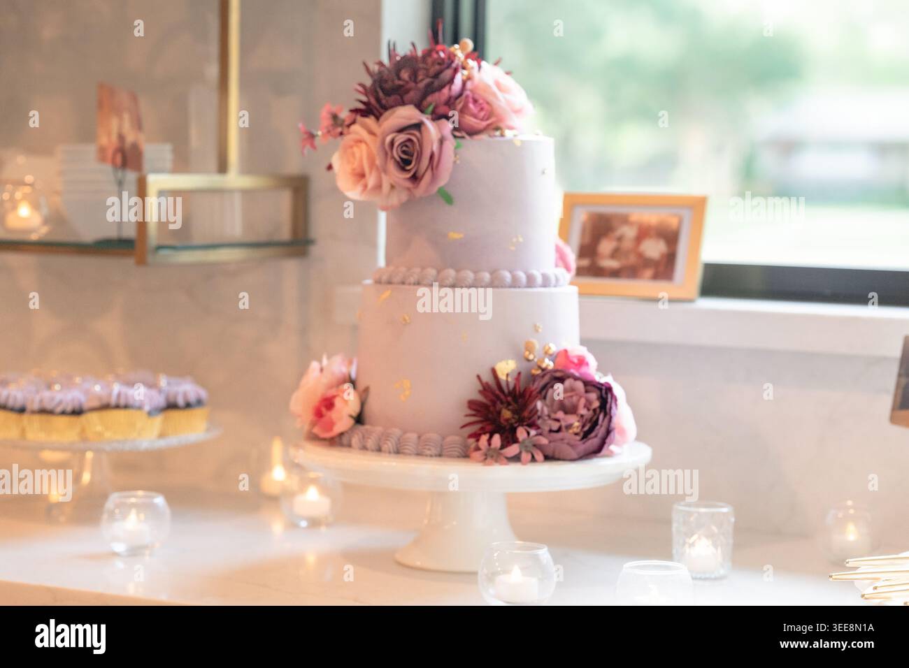 Zwei-stöckiger weißer Festkuchendekorierter mit rosa und burgunderroten Rosen und Dahlien auf einem Kuchenständer, mit glasigen Cupcakes und Kerzenlicht-Votivkerzen Stockfoto